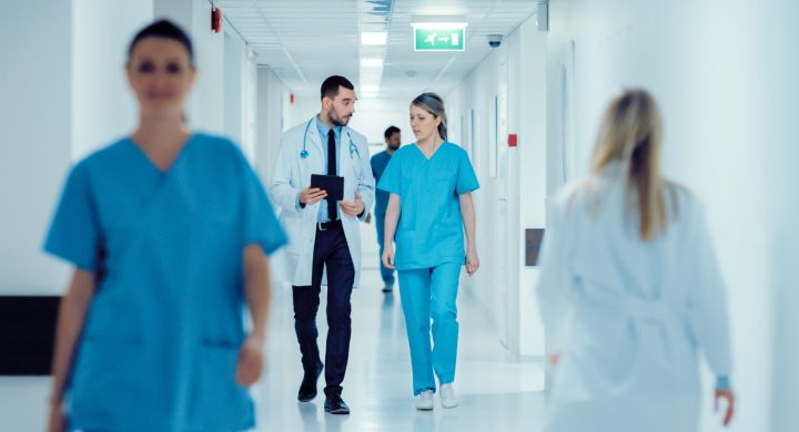 Surgeon and Female Doctor Walk Through Hospital Hallway, They Consult Digital Tablet Computer while Talking about Patient's Health. Modern Bright Hospital with Professional Staff.